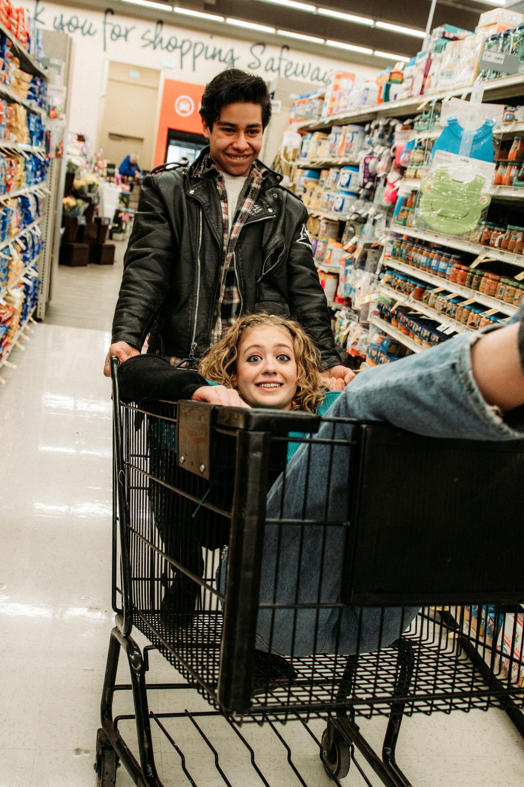 Deux jeunes dans une allée de supermarché, l’un poussant un caddie dans lequel l’autre est assis en riant.